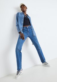 Denim košulja s dugim rukavima, crni uski top, svijetloplave traperice i sive tenisice s prugama. Model pozira u odnosu na bijelu pozadinu.
