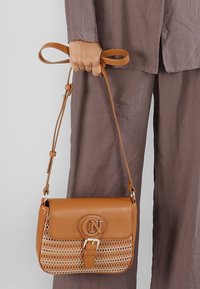 Borsa a tracolla in pelle beige con un motivo intrecciato nella parte inferiore, un logo circolare e una chiusura a fibbia. La tracolla è regolabile.
