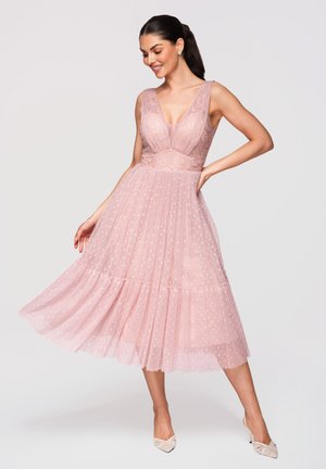 Femme en robe midi rose sans manches en dentelle et tulle à pois, posant d'une main sur la hanche et l'autre tenant la jupe.