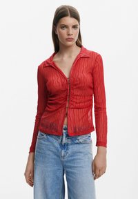 Rotes, durchsichtiges Langarmhemd mit einem strukturierten Wellenmuster, ausgestattet mit einem Kragen und einer Knopfleiste vorne, kombiniert mit hellblauen Jeans.
