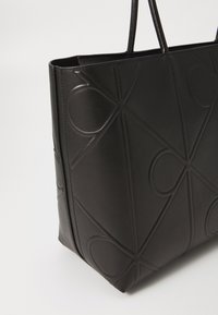 Zwarte leren tote bag met een geperforeerd geometrisch patroon, voorzien van lange hengsels en een gestructureerde vorm. Matte afwerking met een gladde textuur.