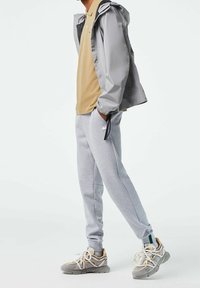 Veste imperméable gris clair avec capuche, polo beige, sweatpants gris et baskets grises avec accents crème et lacets.