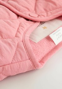 Giacca imbottita per neonati in morbido tessuto rosa, con un'esterno liscio, fodera pelosa, colletto arrotondato e chiusura a pressione. Etichetta visibile all'interno.