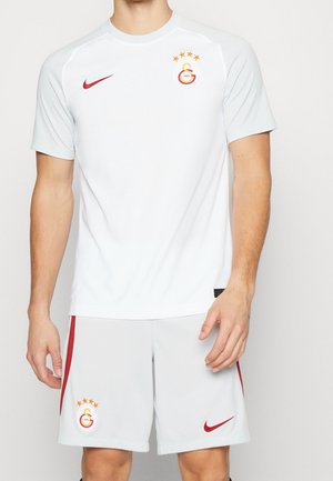 Mann trägt weißes Galatasaray-Fußballtrikot mit roten Nike-Logos und Vereinsabzeichen auf Hemd und Shorts, steht vor einem einfarbigen Hintergrund.