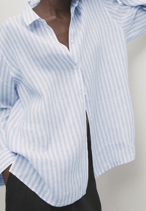 Person wearing a loose light blue and white vertical striped button-up shirt with dark pants, hand partly in pocket.