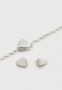 Ασημένιο βραχιόλι σε σχήμα καρδιάς με χάραξη GUESS και διακοσμητικά στρας, συνοδευόμενο από δύο ταίριασμα καρδιάς σκουλαρίκια studs.