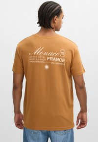 Camiseta de algodón marrón con texto impreso en blanco en la parte trasera, que incluye "Mónaco", "FRANCIA" y "SOCIAL CLUB". Manga corta, corte relajado.