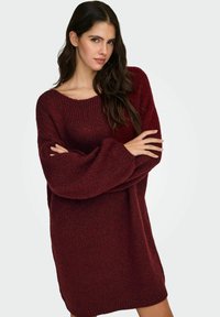 Przesadnie duży bordowy sweter z szerokim dekoltem i fakturowaną tkaniną. Długie rękawy z ściągaczami, sięgające do połowy uda.