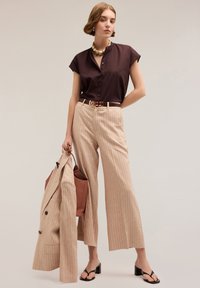 Camicia corta marrone scuro, pantaloni larghi a righe beige, tiene in mano un blazer coordinato e una borsa a tracolla marrone. Sandali neri con tacco.