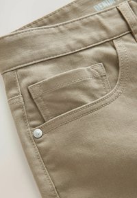 Beige denimbukser med en enkelt baglomme med en sølvknap. Stoffet har en glat tekstur og diskrete syninger.
