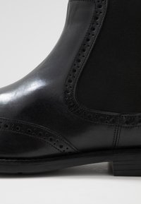 Zwarte leren enkelboot met een gladde afwerking, elastische zijpanelen, decoratieve brogue perforaties en een lage gestapelde hak.