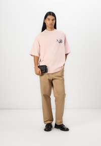 Lys rosa oversized t-skjorte med grafisk design, kombinert med beige bukser og svarte sko. Liten svart crossbody-bag båret på livet.