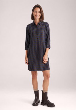 Mujer sonriente que lleva un vestido tipo camisa a cuadros azul y negro con mangas tres cuartos y botines marrón oscuro con calcetines beige.