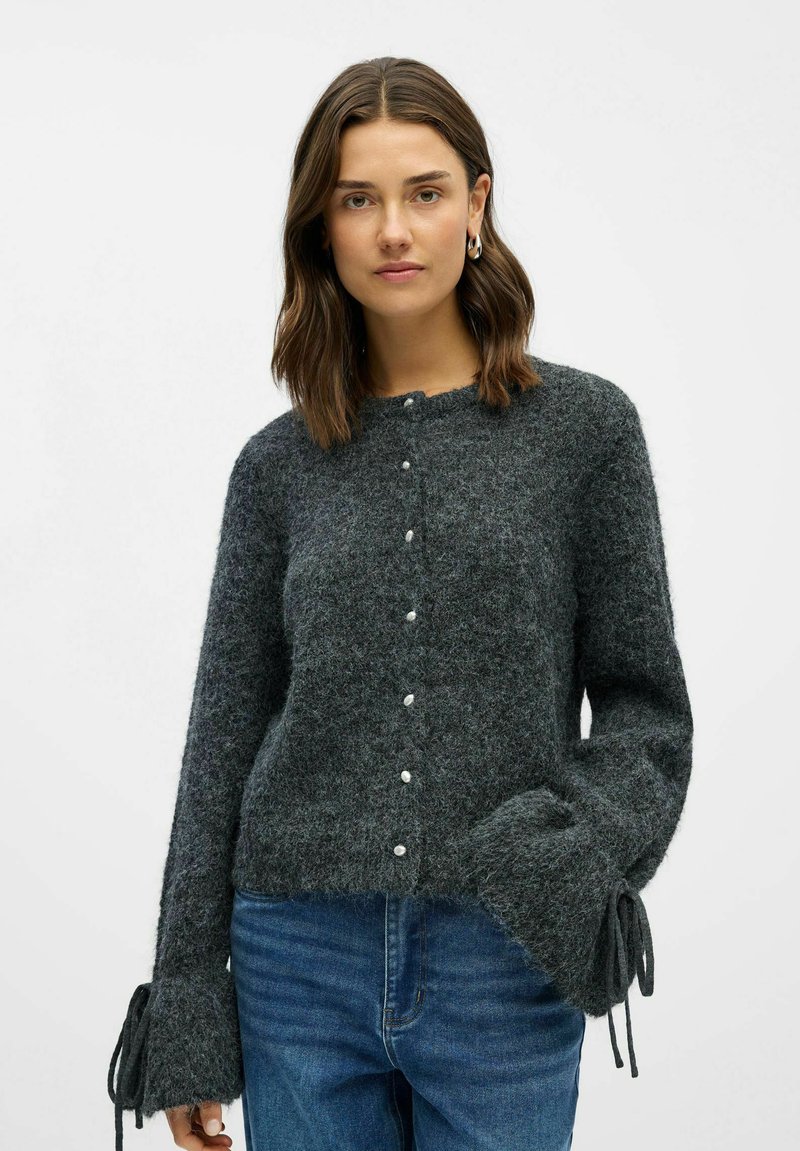 Grå strikket cardigan med lange ærmer, der har tekstureret stof, perleknapper og bundet dekoration ved mansjetterne. Båret sammen med blå jeans.