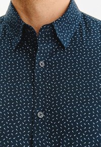 Zbliżenie ciemnoniebieskiej koszuli z guzikami w białe geometryczne wzory oraz kołnierzykiem, noszonej przez osobę z widoczną szyją i podbródkiem.