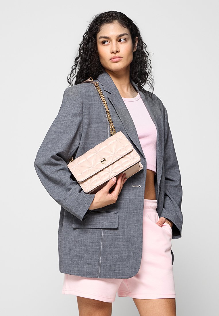 Sac à main en cuir matelassé rose avec une chaîne dorée, tenu contre un blazer gris, présentant une forme rectangulaire et une fermeture à rabat horizontal.