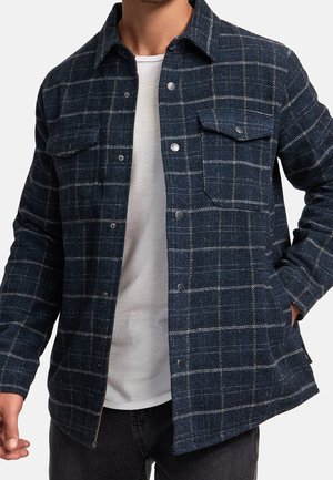 Mann trägt eine dunkelblaue karierte Jacke mit Knöpfen über einem weißen T-Shirt, eine Hand in der Tasche vor einem schlichten Hintergrund.