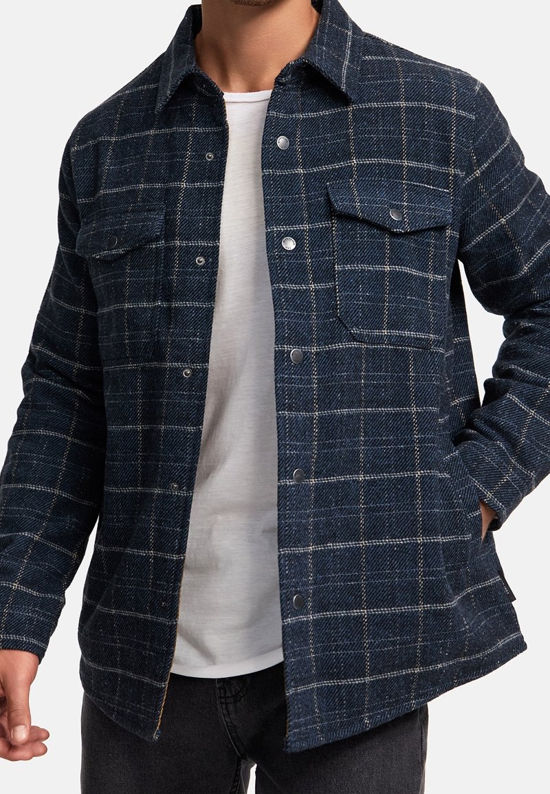 Homme portant une veste à carreaux bleu foncé boutonnée sur un t-shirt blanc, une main dans la poche, sur un fond uni.