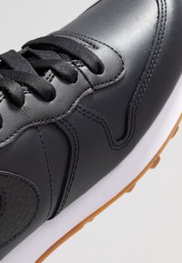 Svart lädersneaker med framträdande sömmar, texturerade detaljer och svarta snören. Vit mellansula och gummisula i brunt fulländar designen.