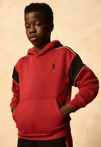 Teenage dreng iført en rød og sort hoodie med hænderne i lommen, ser selvsikker ud mod kameraet mod en beige baggrund.