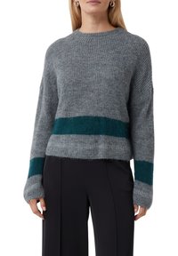 Kobieta w szarej dzianinowej bluzie z ciemnozielonymi poziomymi paskami na rękawach i talii, w zestawieniu z czarnymi spodniami.