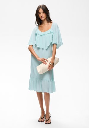 Femme portant une robe bleu clair à volants, tenant une pochette blanche, associée à des sandales noires à brides sur fond blanc.