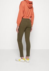 Henkilö, jolla on päällään oliivinvihreät skinny-housut, valkoiset kengät keltaisilla yksityiskohdilla ja lyhyt oranssi huppari, seisoo valkoista taustaa vasten.