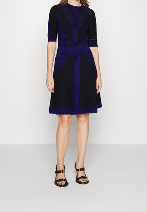 Femme portant une robe noire mi-longue avec des rayures géométriques bleues, manches mi-longues et des sandales noires ouvertes à l’avant avec des brides à la cheville.