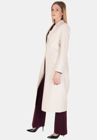 Lungo cappotto color crema con un design su misura, chiusura con un bottone singolo, due tasche frontali, indossato sopra pantaloni bordeaux scuri e tacchi neri.