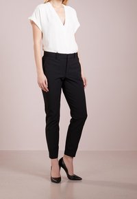 Kvinna iförd en vit kortärmad V-ringad blus, svarta skräddarsydda ankellånga byxor och svarta spetsiga högklackade skor stående på en beige golv.