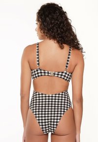 Costume da bagno intero in bianco e nero a quadretti, con spalline regolabili, aperture per le gambe a vita alta e un design con schiena scoperta che presenta una chiusura con clip dorata.