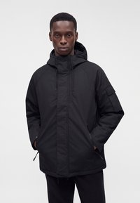 Homme portant une veste à capuche noire, les mains dans les poches, debout devant un fond gris clair uni.
