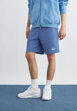 Isik, kes kannab siniseid lühikesi pükse, millel on maailmakuul ja suksu logo, valgeid tossusid, valgeid sokke ja helesinist lukuga kampsunit, seisab sinisel vaibal.