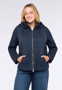 Glunderende jonge vrouw in een marineblauwe gewatteerde hoodie met rits, met haar handen in de zakken en blauwe spijkerbroek tegen een effen achtergrond.