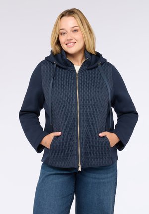 Jeune femme souriante portant un sweat à capuche matelassé bleu marine zippé, les mains dans les poches et un jean bleu, devant un fond uni.