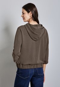 Felpa marrone con cappuccio e vestibilità ampia, a tre quarti di manica, con orlo a coste. Indossata con jeans blu, su uno sfondo chiaro.