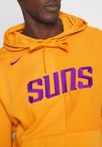 Gros plan sur une personne portant un sweat à capuche orange vif avec le texte "SUNS" en violet et un petit logo Nike violet sur la poitrine.