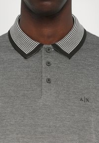 Siva polo majica s prugastim crno-belim ovratnikom, tri gumba na prednjem dijelu i suptilnim logotipom na prsima. Teksturirana tkanina.