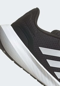 Zapatilla deportiva negra con un upper texturizado, franjas de acento blancas y una suela blanca gruesa y estriada con un capuchón negro en el talón.