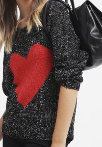 Svart stickad tröja med en stor röd hjärtformad design på framsidan. Ärmarna är lätt uppvikta och Texturen ser mjuk och mysig ut.