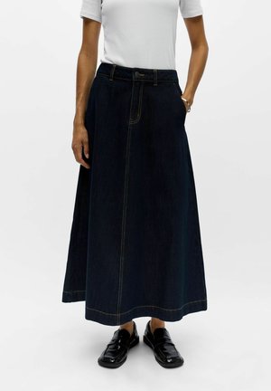 Personne portant un haut blanc à manches courtes, une longue jupe en jean bleu foncé avec une couture devant, et des chaussures noires sans lacets, debout avec une main dans la poche.