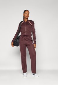 Maroon idrottsoverall med vita ränder, zip-up jacka, byxor med elastisk midja, kombinerad med en svart väska och vita sneakers.