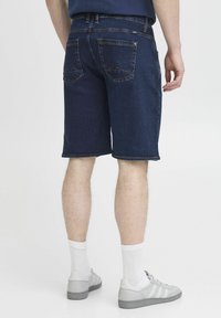 Shorts en denim foncé avec une coupe droite, comportant des poches arrière avec une couture en zigzag discrète. Portés avec des chaussettes blanches et des baskets gris clair.