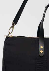 Černá plátěná taška s koženými detaily, obdařená zlatým zipem, zlatými kovovými prvky a texturovaným popruhem. Jednoduchý, funkční design.