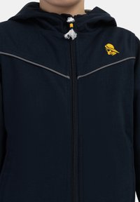 Tamsiai mėlynas užtrauktukas striukė su gobtuvu, kuriame yra šviesiai pilkos juostelės, geltonas logotipas ant krūtinės ir tekstūruotas audinio paviršius.