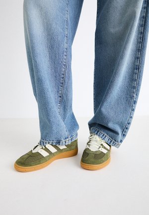 Person wearing light blue wide-leg jeans and green suede sneakers with white stripes and gum soles, standing on a plain white surface.