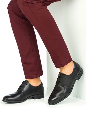 Hombre con zapatos de vestir de cuero negro con cordones y pantalones burdeos, con un pie apoyado contra una pared blanca.