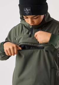 Olivengrøn jakke med en lynlåslomme foran, ribkanter ved ærmerne og en hætte. Sort strikket hue med et logoemblem båret på hovedet.