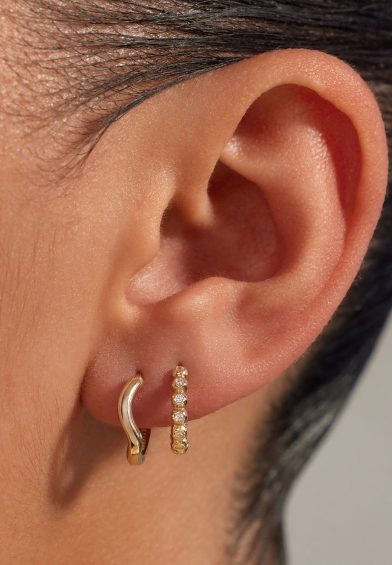 Boucles d'oreilles en or avec un design ondulé et un second petit anneau orné de pierres précieuses transparentes, présentées sur une oreille.