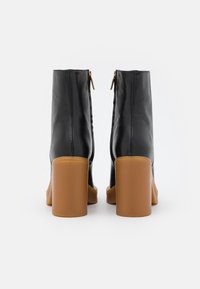 Botas negras de cuero hasta el tobillo con textura lisa, cuentan con tacones gruesos en bloque de color marrón claro y discretas cremalleras doradas en la parte trasera.